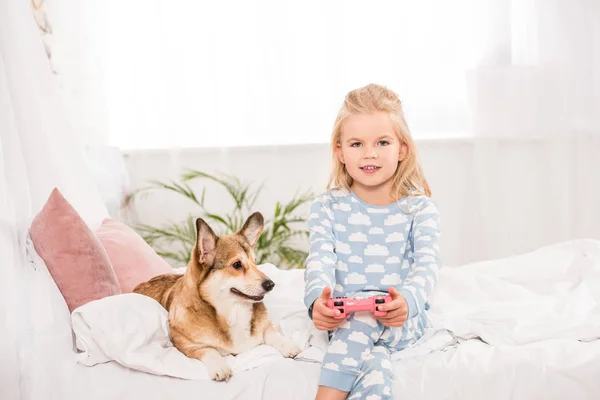 Cute child sitting on bed with pembroke welsh corgi dog, holding joystick and playing video game at home — Stock Photo