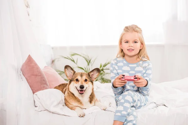 Kind sitzt mit pembroke walisischem Corgi-Hund im Bett, hält Steuerknüppel und spielt zu Hause Videospiel — Stockfoto