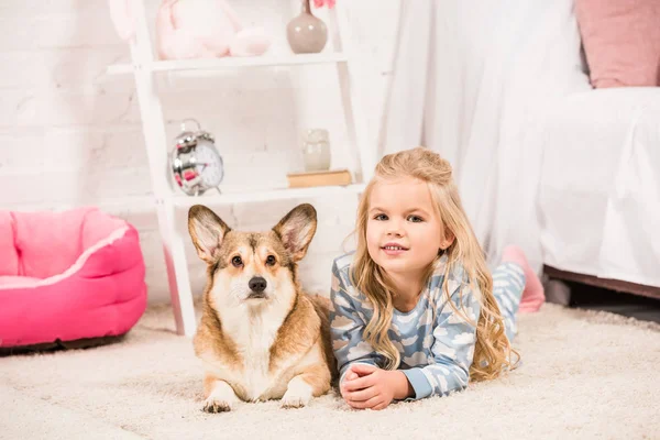 Adorable child in pajamas looking at camera with welsh corgi dog at home — Stock Photo