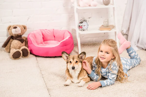 Smiling child in pajamas looking at camera and stroking welsh corgi dog at home — Stock Photo