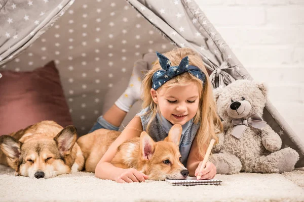 Adorable child lying in wigwam with welsh corgi dogs and writing in notebook at home — Stock Photo