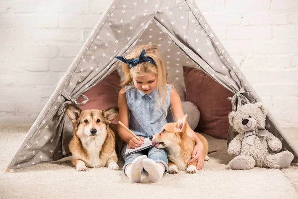 Cute child sitting with corgi dogs in wigwam and writing in notebook at home — Stock Photo