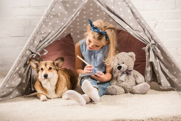 Niedliches Kind sitzt mit Corgi-Hund im Wigwam und schreibt zu Hause in Notizbuch — Stockfoto