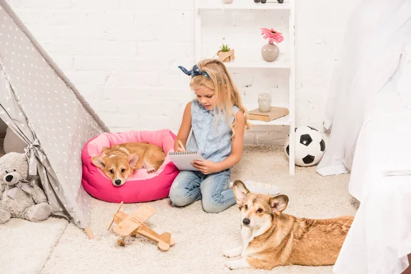 Lindo niño sentado en casa con perros corgi galeses y escritura en cuaderno - foto de stock