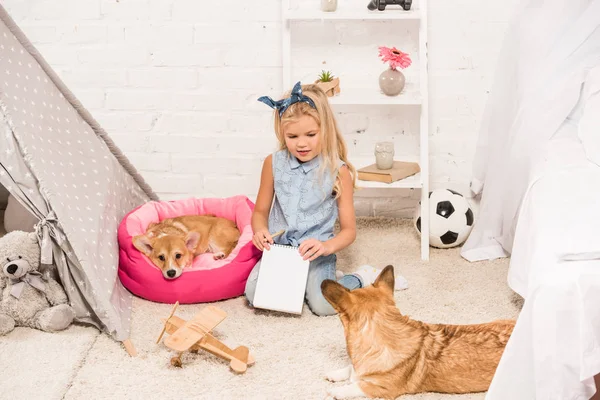 Niedliches Kind sitzt mit welschen Corgi-Hunden zu Hause und hält Notizbuch mit Bleistift in der Hand — Stockfoto