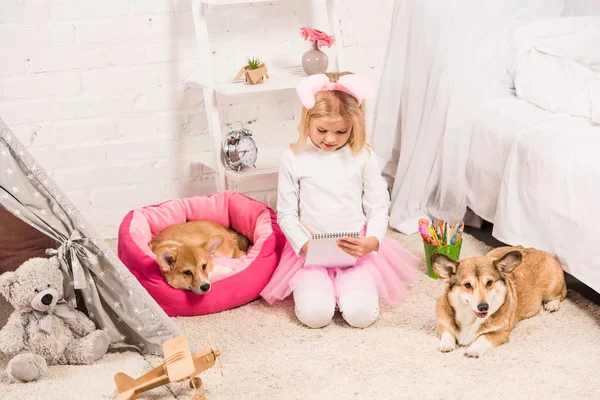 Niedliches Kind mit Hasenohren-Stirnband sitzt mit walisischen Corgi-Hunden zu Hause und schreibt in Notizbuch — Stockfoto