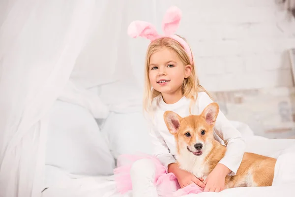 Criança sorridente em orelhas de coelho headband sentado com galês corgi cão na cama em casa — Fotografia de Stock