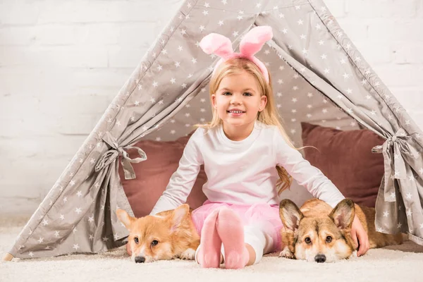 Criança sorridente em orelhas de coelho headband sentado com cães corgi e ursinho de pelúcia em wigwam — Fotografia de Stock