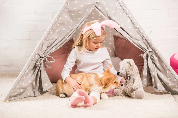 Bambino in fascia orecchie coniglietto seduto con cane corgi e orsacchiotto in wigwam a casa — Foto stock