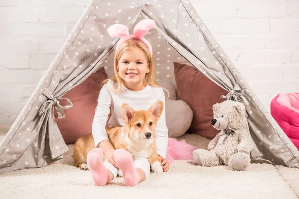 Cute child in bunny ears headband sitting with corgi dog and teddy bear in wigwam — Stock Photo