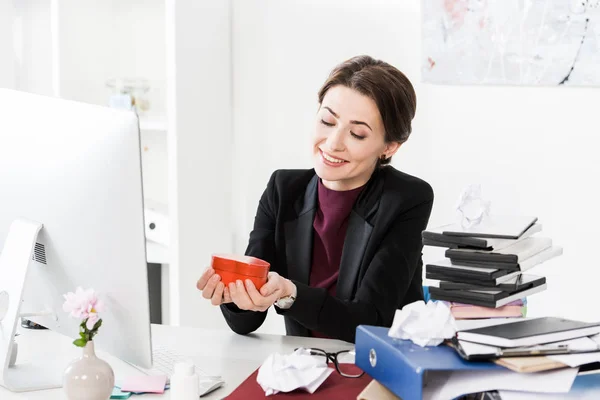 Felice donna d'affari attraente in possesso di cuore a forma di scatola regalo rossa in ufficio — Foto stock