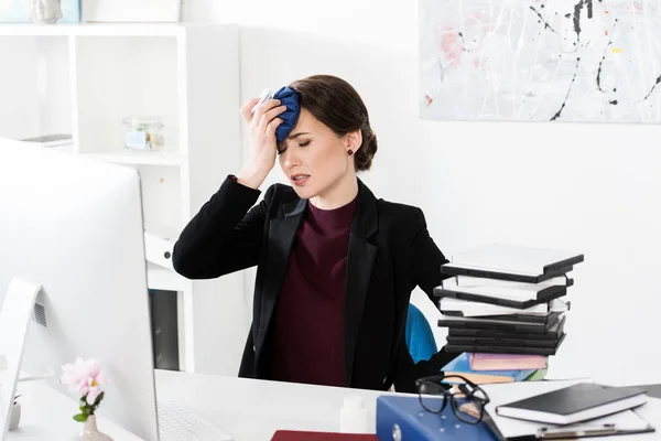Mujer de negocios que tiene dolor de cabeza y tocando la cabeza con bolsa de hielo en la oficina - foto de stock