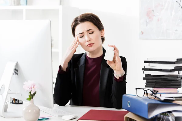Mulher de negócios atraente tendo dor de cabeça e segurando garrafa com pílulas no escritório — Fotografia de Stock