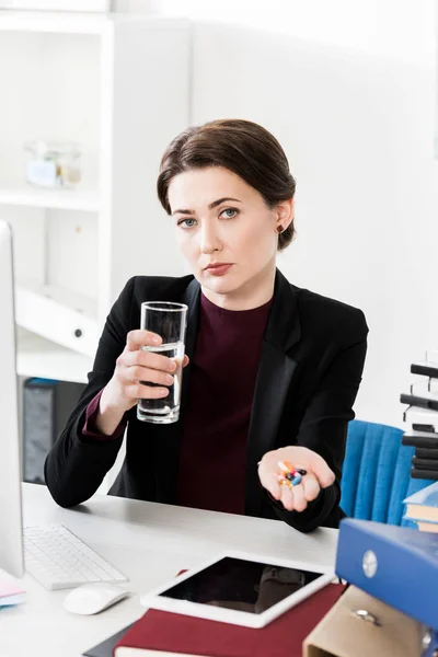 Attraktive Geschäftsfrau, die im Büro ein Glas Wasser und Pillen in die Kamera hält — Stockfoto