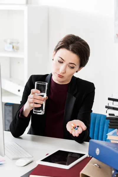 Attraktive Geschäftsfrau mit Glas Wasser und Pillen im Büro — Stockfoto