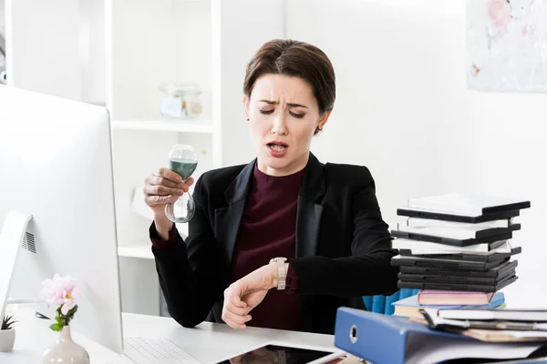 Atractiva mujer de negocios sorprendida sosteniendo reloj de arena y comprobando el tiempo con reloj de pulsera en la oficina - foto de stock