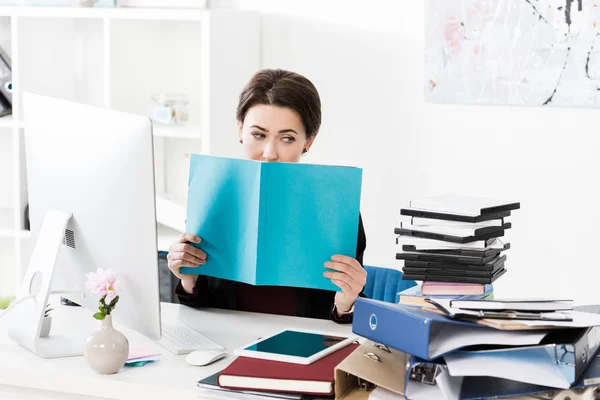 Привлекательная деловая женщина, выглядывающая из бирюзовой книги в офисе — стоковое фото