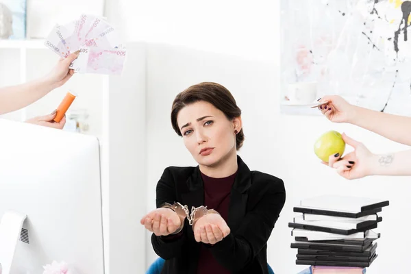 Mulher de negócios triste mostrando as mãos com algemas enquanto secretários segurando dinheiro, xícara e maçã no escritório — Fotografia de Stock