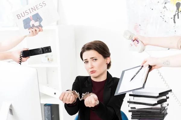 Triste mujer de negocios mostrando las manos con esposas mientras secretarias sosteniendo periódico, teléfonos y portapapeles en la oficina - foto de stock
