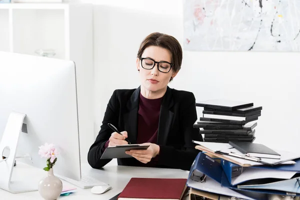 Atractiva mujer de negocios en gafas de escribir algo al portapapeles en la oficina - foto de stock