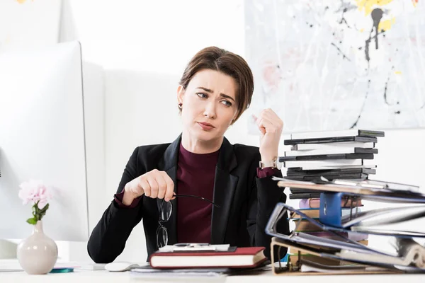 Sad tired attractive businesswoman holding glasses and looking at stack of documents on table in office — Stock Photo