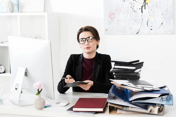 Attraktive Geschäftsfrau mit Brille, Klemmbrett haltend und im Büro wegschauend — Stockfoto