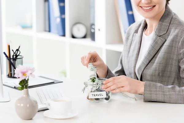 Imagem cortada de empresária feliz colocando dinheiro em frasco de vidro para viagens no escritório — Fotografia de Stock