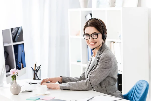 Smiling attractive call center operator in glasses and headset using computer at table in office, looking at camera — Stock Photo