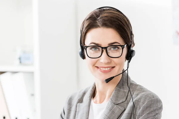 Smiling beautiful call center operator in glasses and headset looking at camera in office — Stock Photo