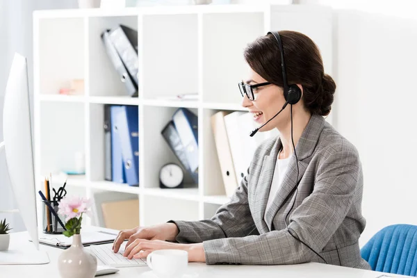 Vista laterale del sorriso attraente operatore call center in occhiali e auricolari utilizzando il computer a tavola in ufficio — Foto stock