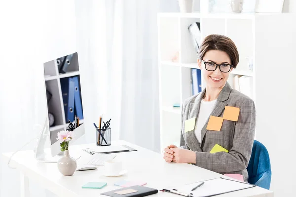 Souriant attrayant femme d'affaires avec des autocollants en papier sur la veste en regardant la caméra dans le bureau — Photo de stock