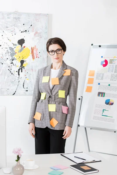 Attraktive Geschäftsfrau mit Papieraufklebern auf Jacke, die neben Flipchart steht und im Büro in die Kamera schaut — Stockfoto