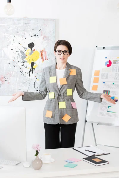 Shocked businesswoman with paper stickers on jacket standing near flipchart and showing shrug gesture in office — Stock Photo