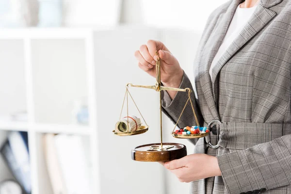 Imagem cortada de advogado segurando escalas de justiça com dinheiro e drogas na mesa no escritório — Fotografia de Stock