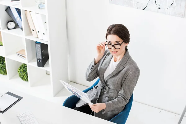 Vista de ángulo alto de atractiva mujer de negocios en gafas con periódico en la oficina y mirando a la cámara - foto de stock