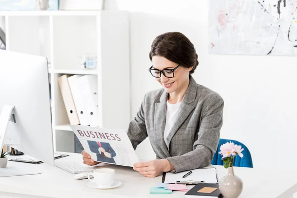 Smiling attractive businesswoman in glasses reading newspaper in office — Stock Photo