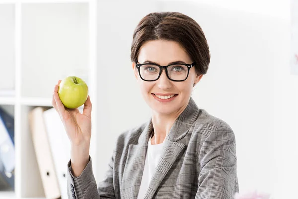 Porträt einer attraktiven Geschäftsfrau, die einen Apfel im Büro hält und in die Kamera blickt — Stockfoto