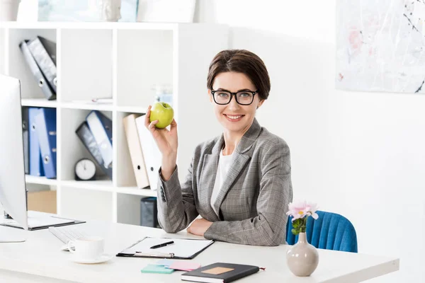 Улыбающаяся привлекательная деловая женщина, держащая яблоко в офисе и смотрящая в камеру — стоковое фото