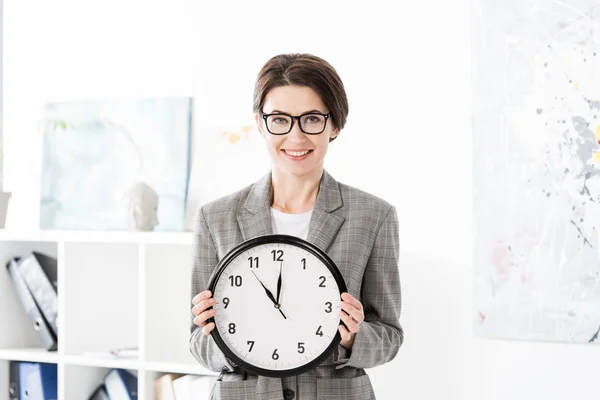 Retrato de mulher de negócios atraente sorridente segurando relógio de parede e olhando para a câmera no escritório — Fotografia de Stock