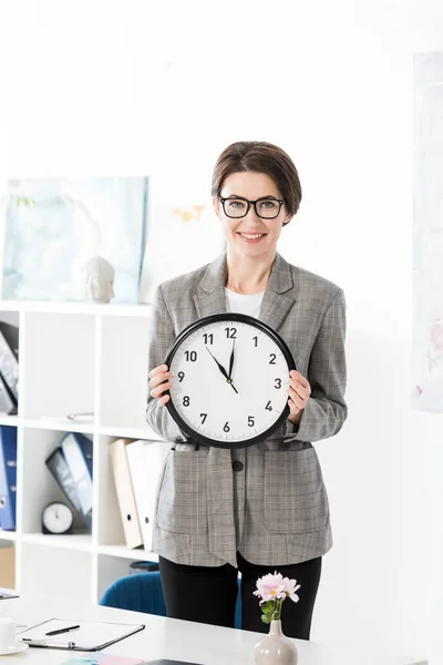 Улыбающаяся красивая деловая женщина, держащая настенные часы и смотрящая на камеру в офисе — стоковое фото