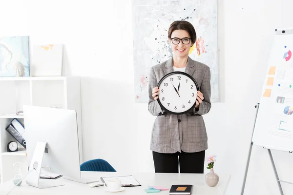 Sonriente atractiva mujer de negocios sosteniendo reloj de pared y mirando a la cámara en la oficina - foto de stock