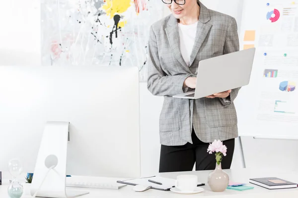Imagen recortada de la mujer de negocios sonriente sosteniendo el ordenador portátil en la oficina - foto de stock