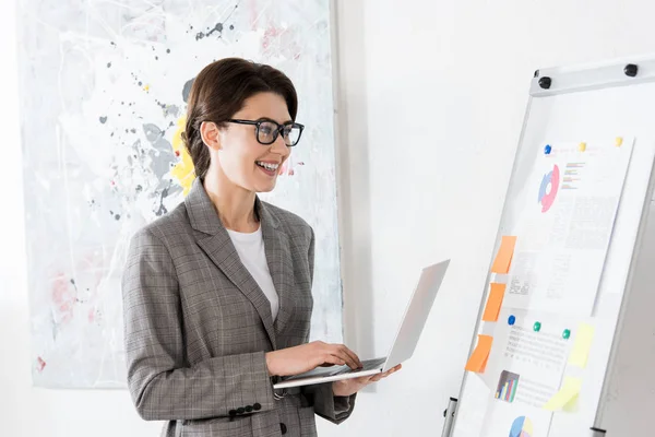 Smiling attractive businesswoman using laptop and looking at flipchart in office — Stock Photo