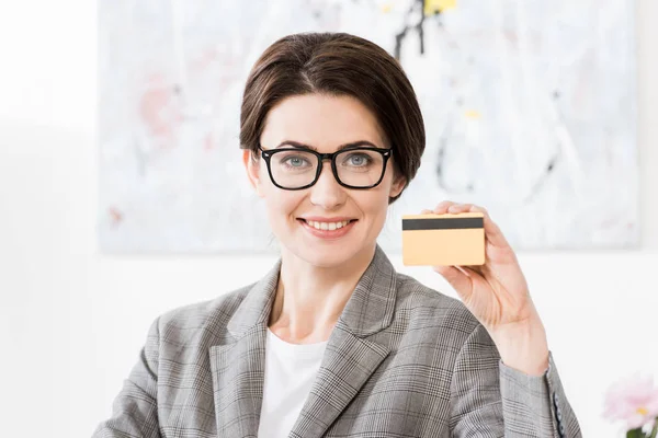 Attractive businesswoman in glasses showing credit card and looking at camera in office — Stock Photo