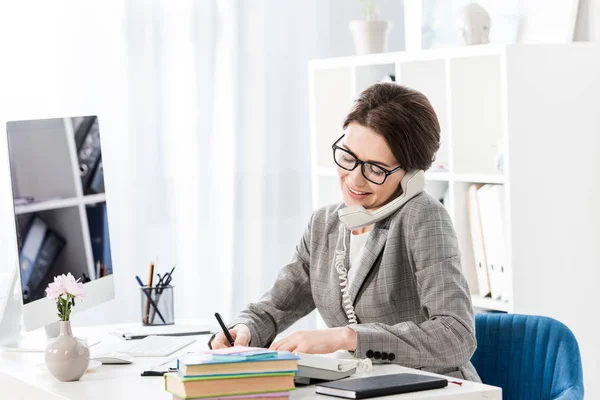 Улыбающаяся привлекательная деловая женщина разговаривает по стационарному телефону в офисе — стоковое фото