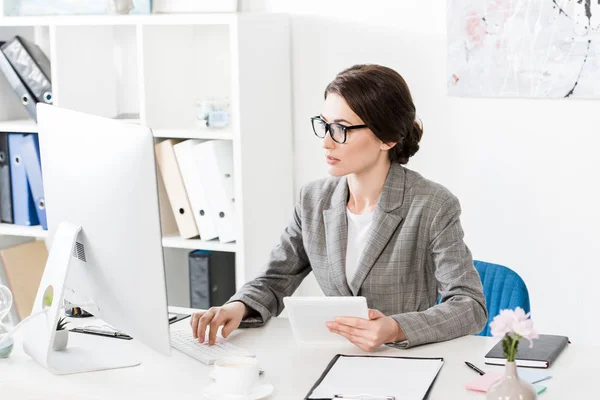 Séduisante femme d'affaires en costume gris tenant tablette et regardant l'ordinateur dans le bureau — Photo de stock