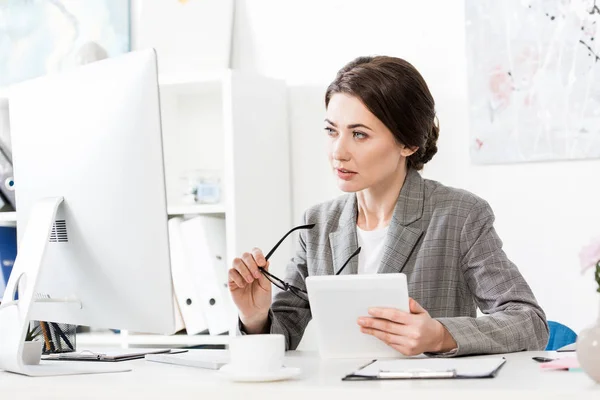 Красива бізнес-леді в сірому костюмі тримає планшет і дивиться на комп'ютер в офісі — стокове фото