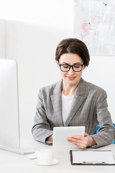 Alegre atractiva mujer de negocios en traje gris usando tableta en la oficina - foto de stock