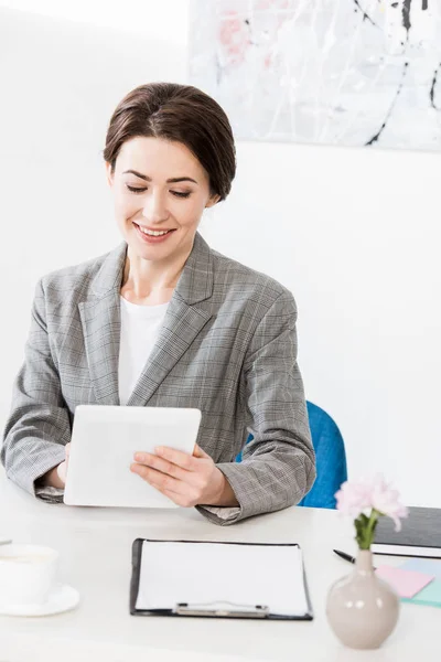 Donna d'affari attraente sorridente in abito grigio utilizzando tablet in ufficio — Foto stock