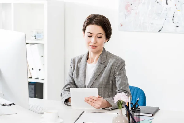 Atractiva mujer de negocios en traje gris usando tableta en la mesa en la oficina - foto de stock